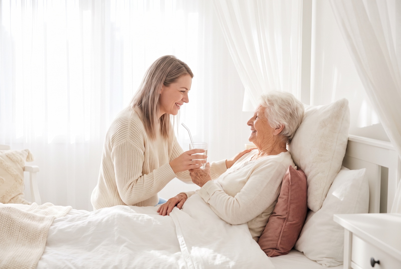 Betreuungskraft reicht bettlägeriger Seniorin ein Glas Wasser