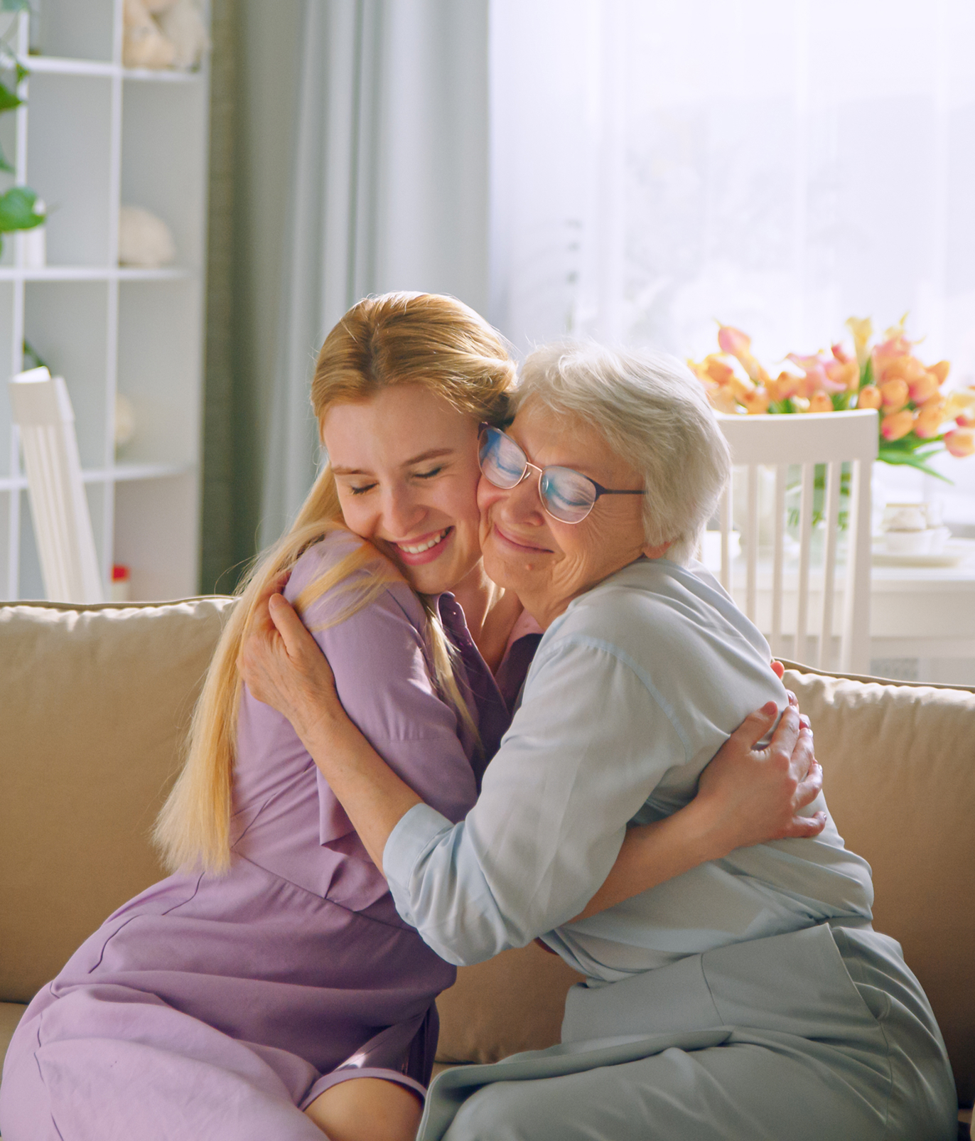 Junge Frau umarmt eine lachende Seniorin auf dem Sofa – Seniorenbetreuung zu Hause in Prien am Chiemsee